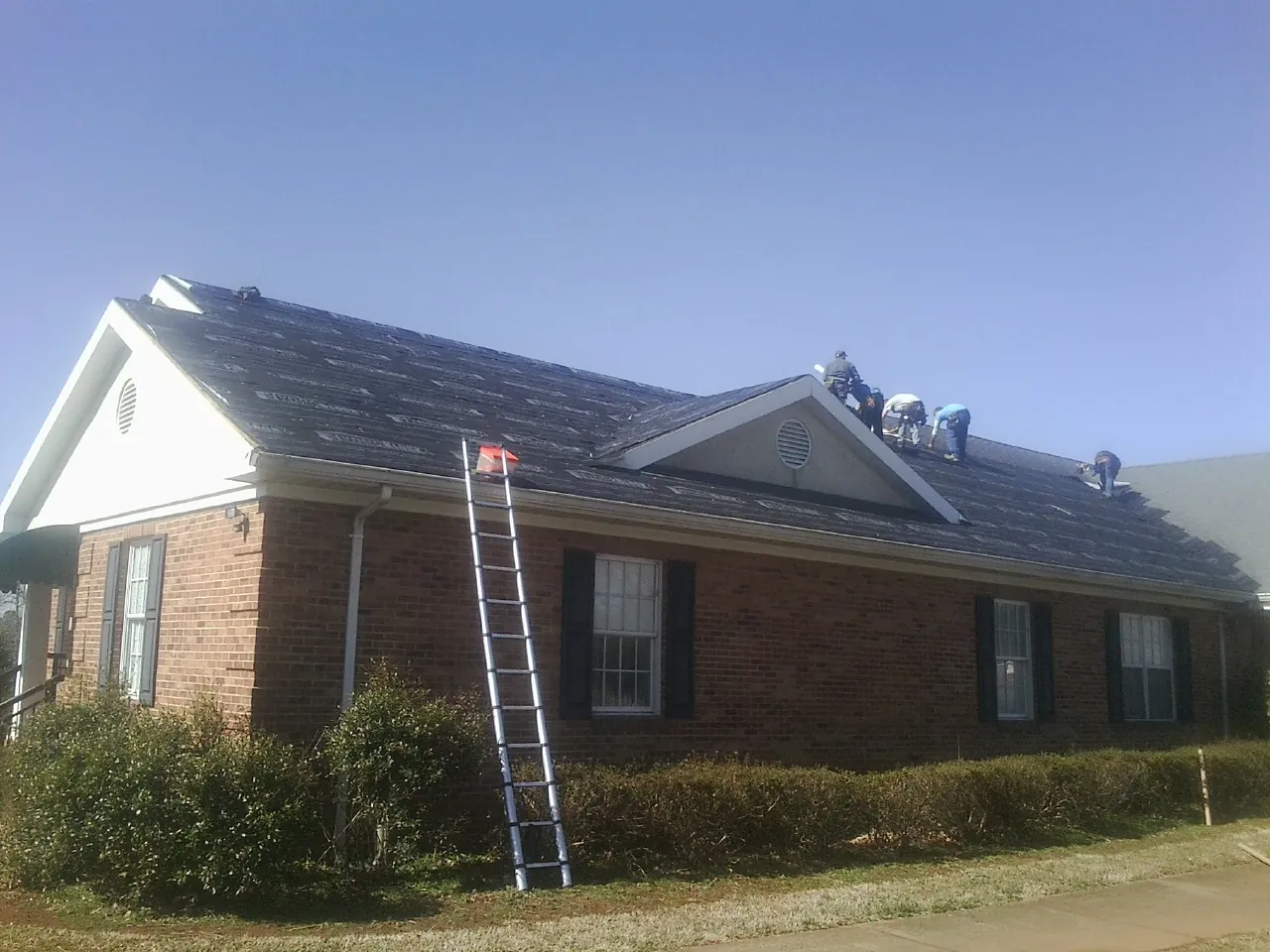 Crew tearing shingles off a brick ranch roof with a ladder set — Metal Roof Repair work by our crew in Mc Coll