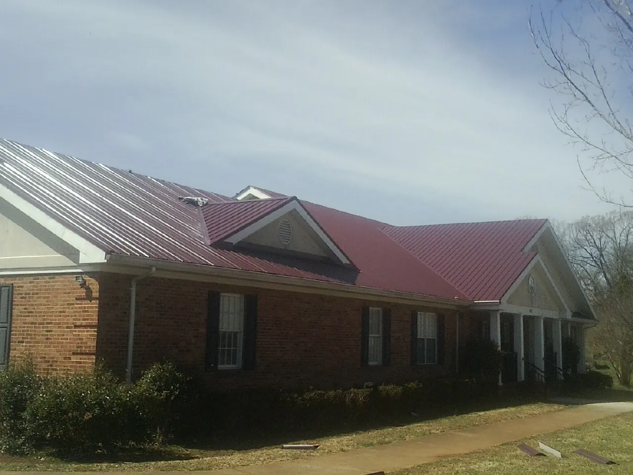 Red metal roof with gabled entries on a long brick building, from a Hail Damage Roof Repair job in Mc Coll