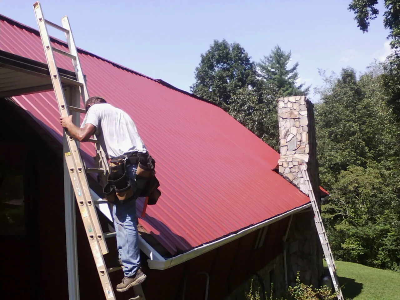 Roofer on a ladder beside a red metal roof and stone chimney — Soffit & Fascia Repair by Roofing Brothers in Mc Coll